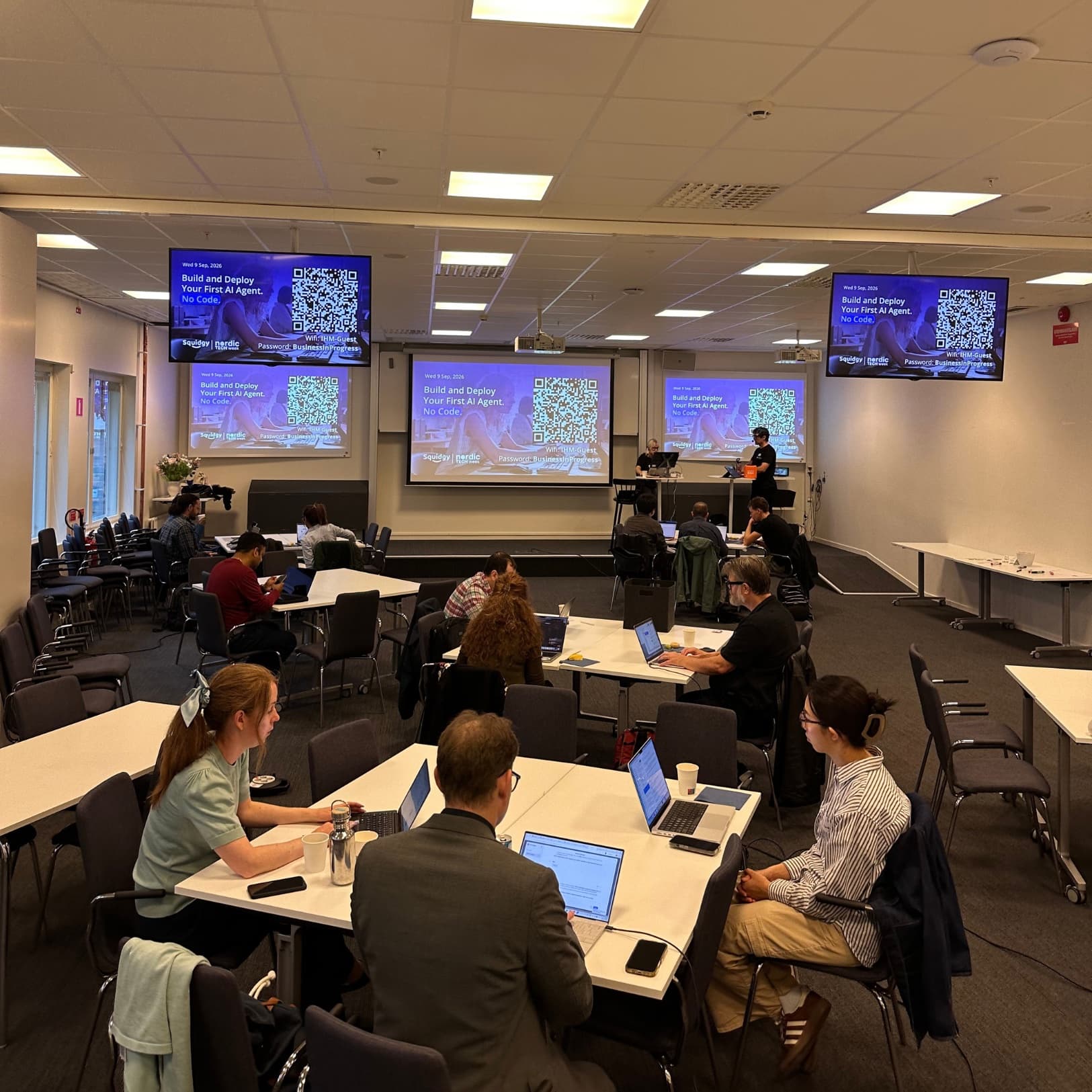 Attendees working on laptops at tables in a workshop room, with screens reading Build and Deploy Your First AI Agent. No Code.